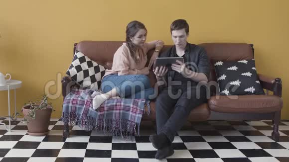 年轻的微笑夫妇与数字平板放松在沙发在家里时尚的房间库存录像男人和女人浏览视频的预览图