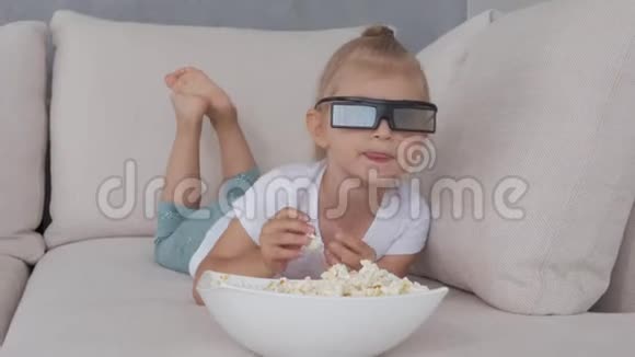 一个小女孩在柔软的沙发上吃爆米花看着3D眼镜里的电视屏幕视频的预览图