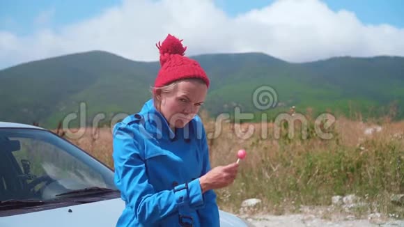 有趣的女孩站在阳光明媚的夏天金发女郎很有趣穿着蓝色外套戴着红色帽子舔着棒棒糖视频的预览图