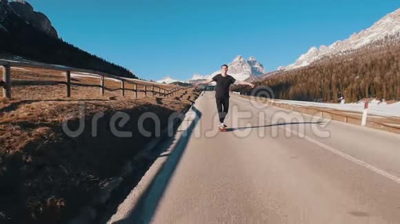 年轻人骑在马路上在森林和山脉的背景下用一个小滑板保持平衡视频的预览图