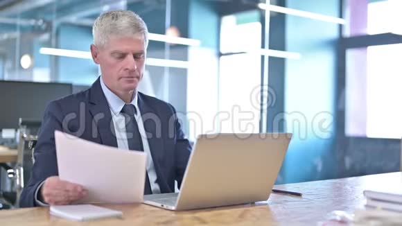 中年商人阅读文件和使用笔记本电脑视频的预览图