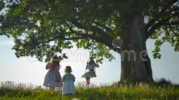两个女儿沿着绿地向母亲走去在绑在老树上的秋千上荡秋千视频的预览图