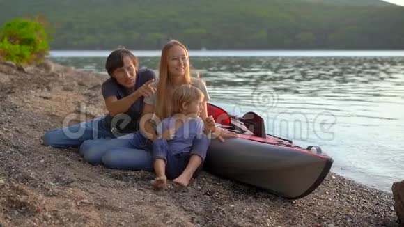一个年轻的家庭父亲母亲和儿子坐在湖边或海滨的充气皮艇旁慢镜头视频的预览图