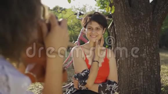 有短发的年轻女子坐在树下而她的朋友在花园里拍照自行车站着视频的预览图