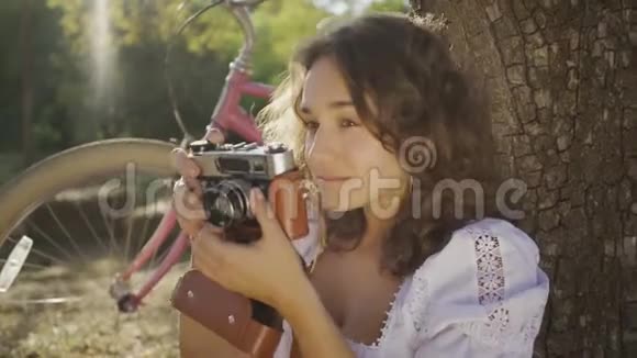迷人的年轻摄影师卷发坐在树下在花园或公园里用旧相机拍照视频的预览图