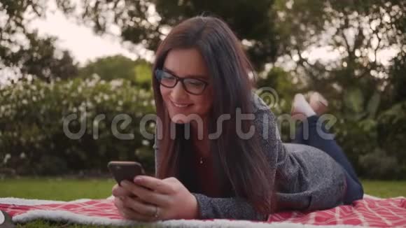 使用智能手机躺在公园的毯子上轻松微笑的年轻女子的肖像视频的预览图