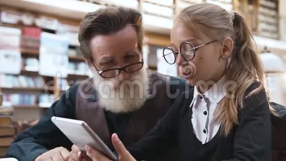 英俊的老胡子祖父带着眼镜和他微笑的十几岁的孙女说话在图书馆里用电子书视频的预览图