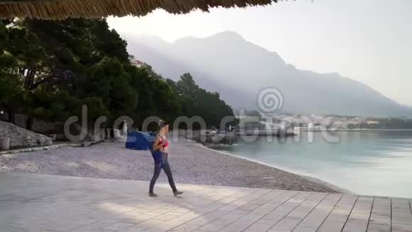 穿着运动服带瑜伽垫的赤脚女人走在海边的石板上视频的预览图