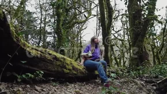 一个穿着休闲风格的快乐女孩坐在长满苔藓的树上抬头望着一片石树林视频的预览图