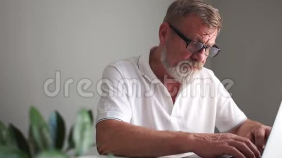 一个留着眼镜和白色T恤的胡子的人在笔记本上记笔记并与笔记本电脑一起工作商业和视频的预览图