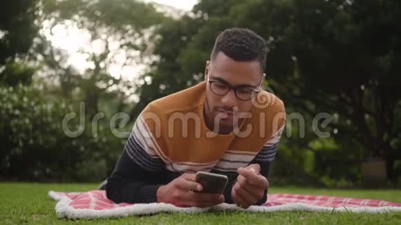一位微笑的非洲年轻人躺在草地上的毯子上在公园的手机上自拍的肖像视频的预览图