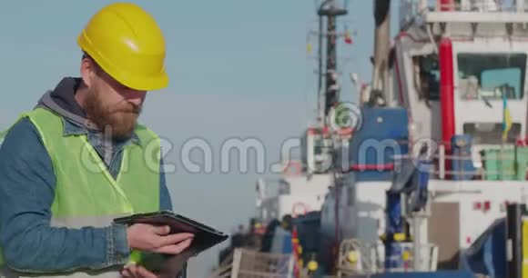 港口工人与一个胡子在一个黄色头盔立场与一个平板电脑在海港与船的背景码头工人视频的预览图