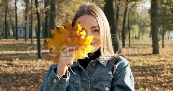美丽的微笑的女人穿着牛仔裤夹克在黄色的秋天公园里她脸上挂着一片落叶视频的预览图