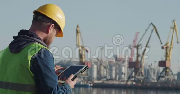 一个在黄色头盔中有胡子的港口工人在海岸的背景下使用平板电脑领班视频的预览图