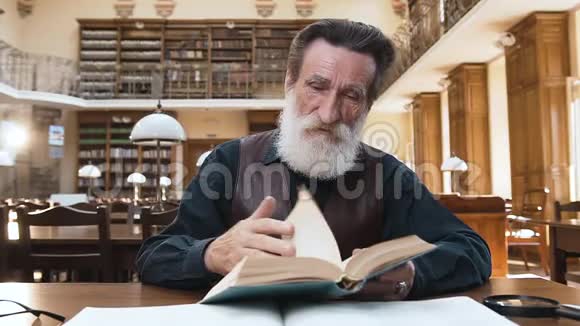 坐在图书馆桌子旁翻书页的一个专注的大胡子老头视频的预览图