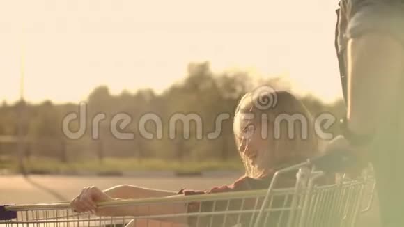 后景快乐的年轻情侣恋爱中的男人和女人在超市停车场的推车里嬉笑打闹视频的预览图