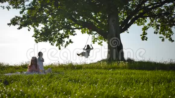 美丽的女人穿着一件乡村白色连衣裙在秋千上摆动她的女儿在田野里视频的预览图