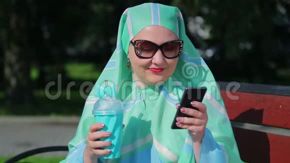 一位戴着浅色围巾和墨镜的年轻穆斯林妇女坐在公园的长凳上喝着咖啡在公园里聊天视频的预览图