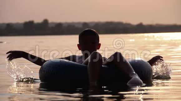 日落时分欢乐男孩在河中的充气圈上游泳的剪影慢动作视频的预览图