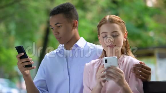 拥抱公园里坐在长椅上的多民族青少年夫妇聊天的智能手机视频的预览图