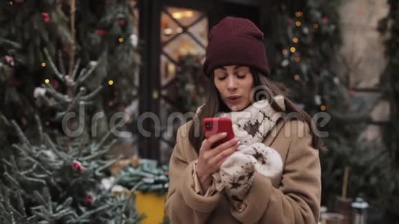 戴着冬帽和手套的漂亮女孩用智能手机聊天看着屏幕看上去既惊讶又惊讶视频的预览图