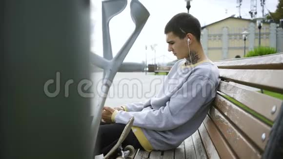 帅哥坐在公园的长凳上用手机听音乐拐杖和滑板就在附近视频的预览图