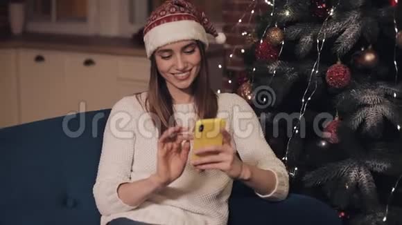 美丽微笑的年轻女士戴着桑塔帽坐在圣诞树附近的沙发上使用她的智能手机概念视频的预览图