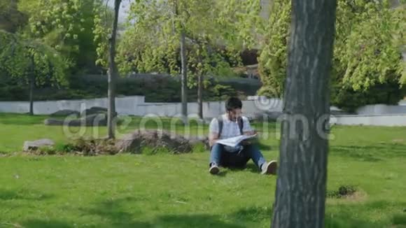 戴眼镜阅读摘要的印度学生坐在公园的草坪上视频的预览图