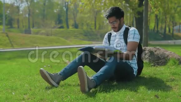 戴眼镜的印度学生带着背包看着坐在城市公园草坪上的抽象物视频的预览图