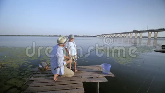 欢乐的消遣父亲和儿子戴着草帽在家庭夏天的天空和河流的背景下快乐地在码头上捕鱼视频的预览图