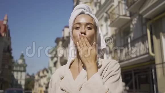 微笑的年轻女子头上戴着毛巾的肖像在站在街上时用空气亲吻看镜头视频的预览图
