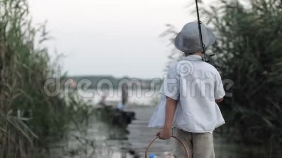 快乐的童年带着钓竿和篮子吃零食的漂亮小男孩沿着桥走到芦苇丛中的码头去钓鱼视频的预览图