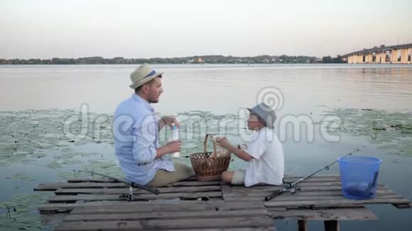 在河边野餐爸爸和儿子钓完鱼后坐在河边的码头上在芦苇丛中享受社交和度假的乐趣视频的预览图
