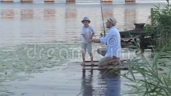 家庭周末父亲和他的小儿子在靠近水的码头上钓鱼后沿着木板上岸视频的预览图
