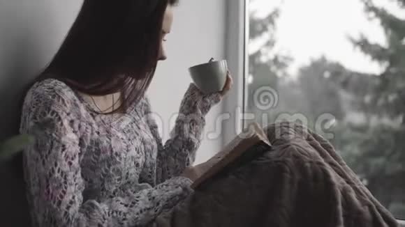 漂亮的年轻女士正坐在窗台上看书在房子里喝茶大窗户绿植视频的预览图