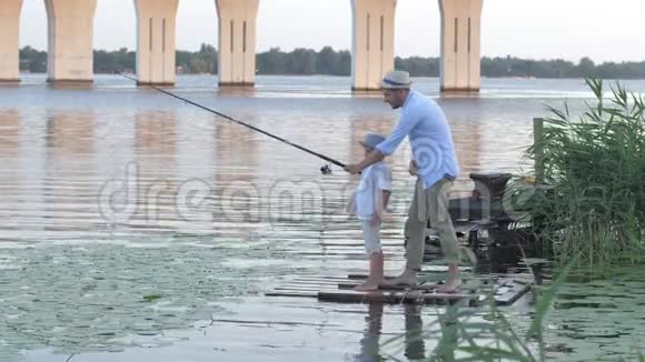 家庭日渔夫父亲周末在河边的芦苇丛中教他的小儿子在码头上钓鱼视频的预览图