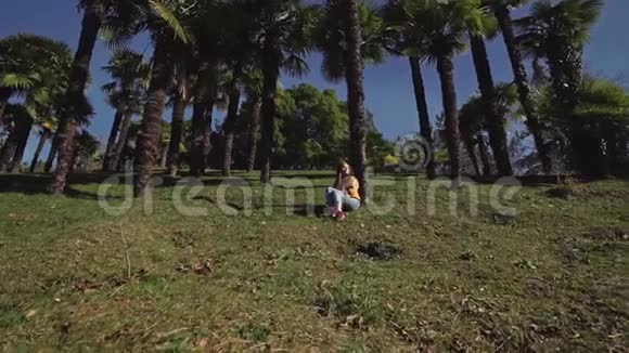 穿着牛仔裤和夹克的漂亮女孩坐在棕榈树下的绿色草坪上视频的预览图