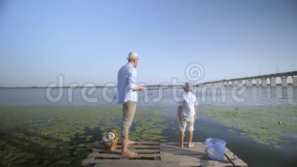 快乐的周末爸爸和儿子戴着太阳帽在码头附近的水上拿着鱼竿爸爸把鱼从河里捞出来视频的预览图