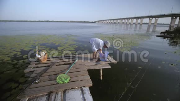 小渔夫戴着帽子的小快乐的孩子从他在河边的码头上钓到的塑料桶鱼拿出来视频的预览图