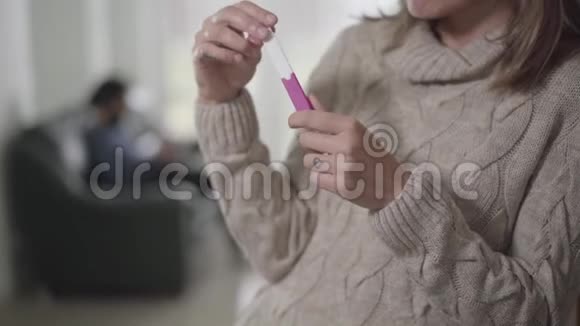 年轻的白种人女人拿着怀孕测试当模糊的男人坐在沙发上的背景妻子在路上思考视频的预览图