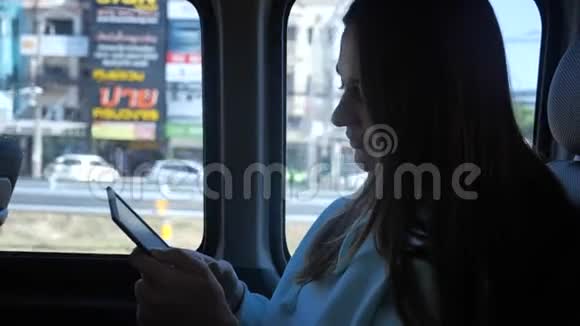 一个年轻的女人正在公共汽车上读一本电子书在窗外阳光明媚的一天背景上的棕榈树4k视频的预览图