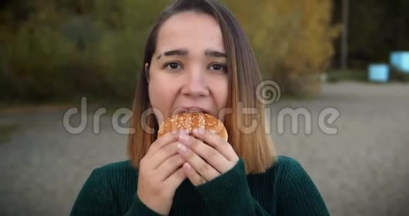 穿着绿色毛衣在大自然的背景下吃大汉堡的知足女孩女孩在大自然中咬一个汉堡视频的预览图