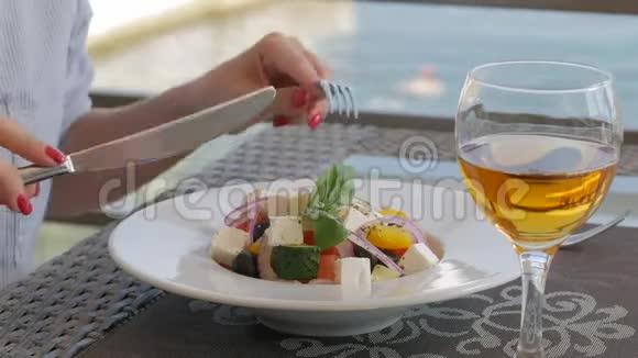 一个拿着刀叉的女人在海滩上的一家餐馆里吃一份漂亮的希腊沙拉沙拉盘的特写视频的预览图