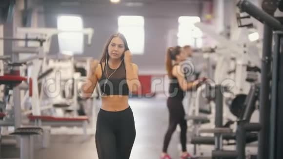 一位运动员女士在健身房里散步戴上耳机把手伸进手柄开始训练视频的预览图