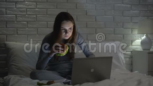 女人咬着美味可口的汉堡晚上在床上吃垃圾食品视频的预览图