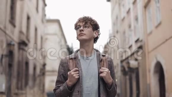 戴眼镜的年轻嬉皮士穿着带包走的耳机里的带支票夹克环顾老城街视频的预览图
