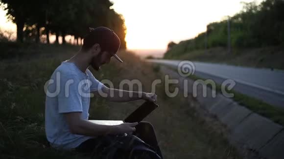 年轻人坐在草地上在沥青路附近的日落时使用笔记本电脑视频的预览图