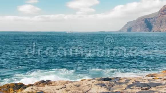 孤独的游艇在海上航行背景上有巨大的岩石黄色岩石沸腾的绿松石冲浪正午耀眼的太阳视频的预览图