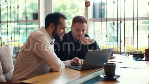 两个年轻的商人坐在桌子旁靠窗放着笔记本电脑的画像视频的预览图