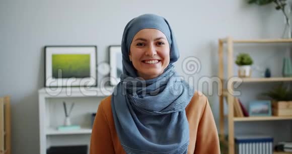 戴着头巾的快乐穆斯林女士在公寓里对着摄像机微笑的照片视频的预览图